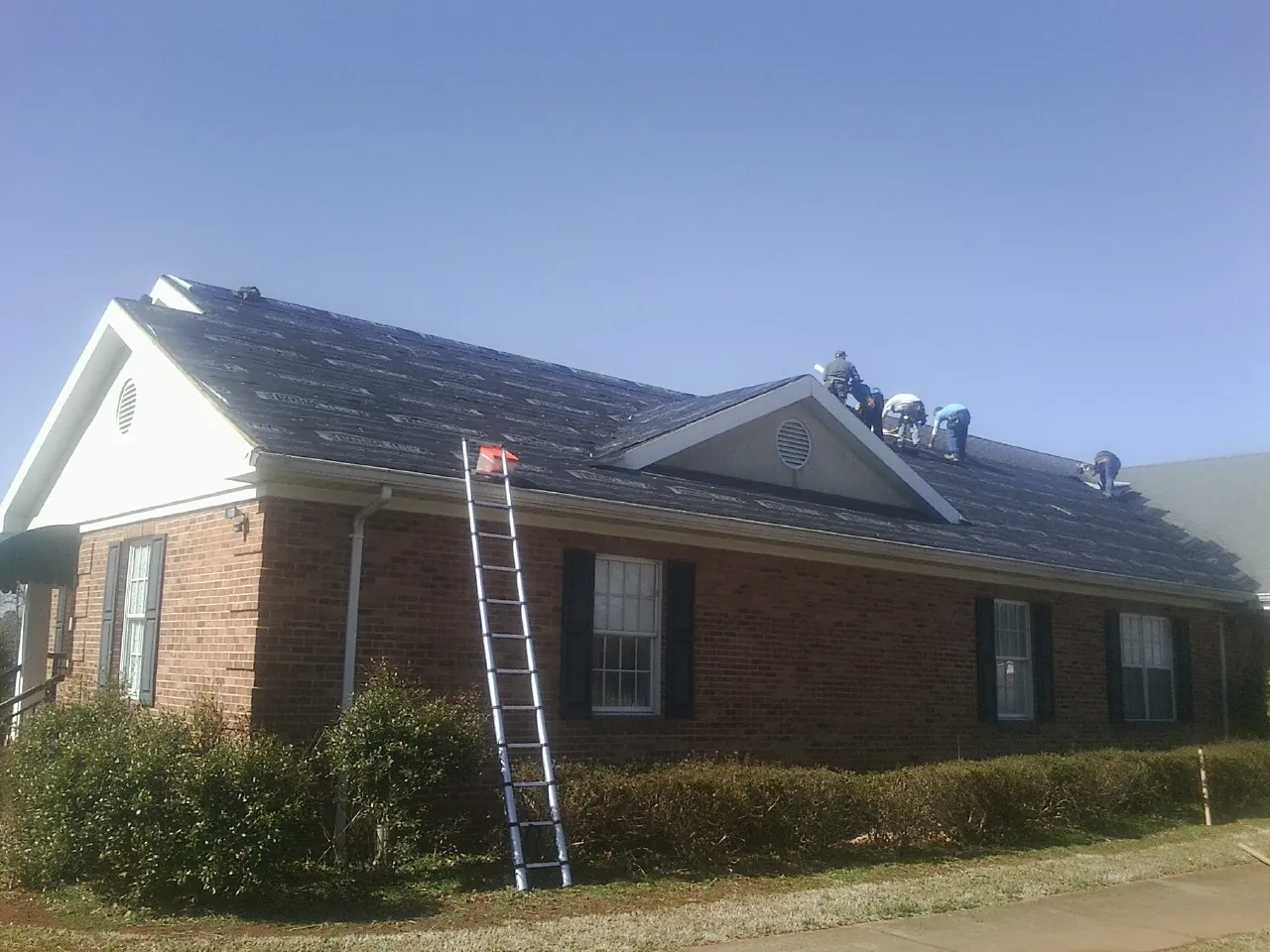 Crew tearing shingles off a brick ranch roof with a ladder set — Emergency Roof Repair work by our crew in Bedford